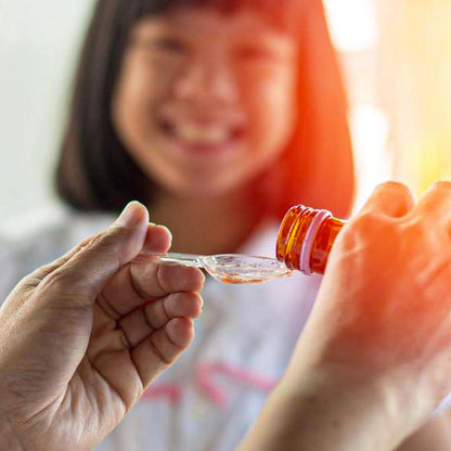 Child smiling while receiving syrup from a bottle, showcasing a caring moment in a family setting.